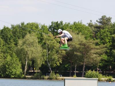 Persona con casco salta en wakeboard sobre el agua con fondo boscoso.