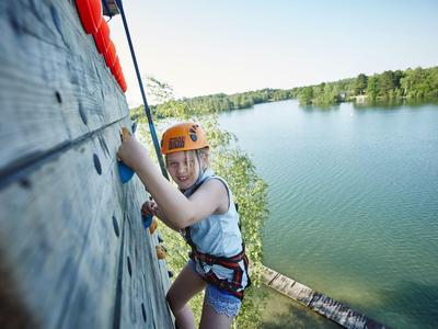 Persona escalando un muro con lago y árboles al fondo.