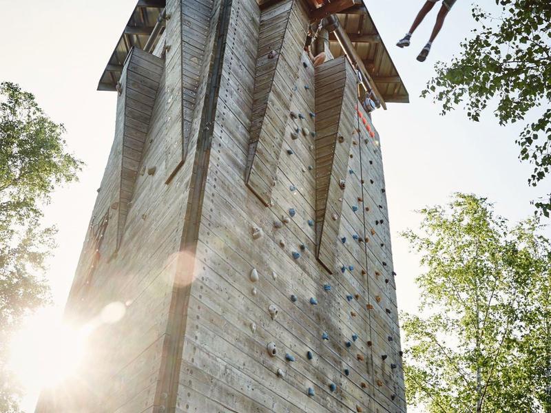 Torre de escalada alta al aire libre con una persona columpiándose desde la cima en un entorno natural