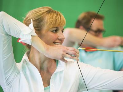 Frau übt Bogenschießen drinnen in Sportkleidung mit grünem Hintergrund.