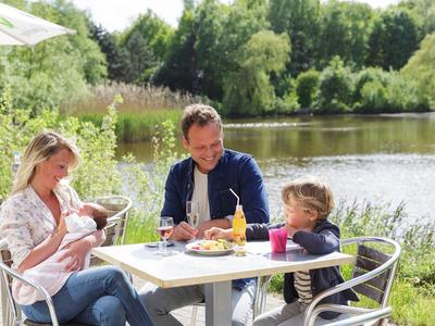 Familie sitzt an einem Tisch im Freien neben einem See und genießt eine Mahlzeit.