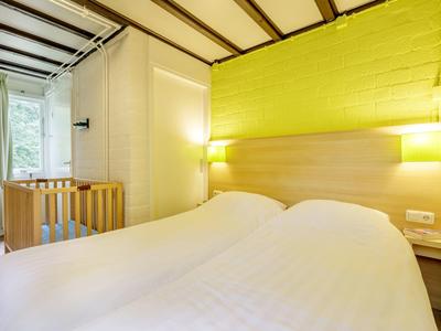 Przytulny pokój hotelowy z podwójnym łóżkiem, żółtą ścianą akcentową i łóżeczkiem dziecięcym przy oknie.