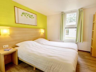 Jasny pokój hotelowy z łóżkiem podwójnym, drewnianą podłogą i zielonym akcentem na ścianie.