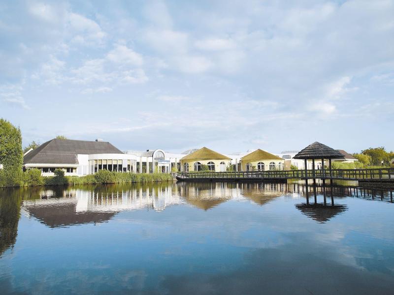 Budynki hotelu z drewnianym molo i altaną odbijające się w spokojnej wodzie.