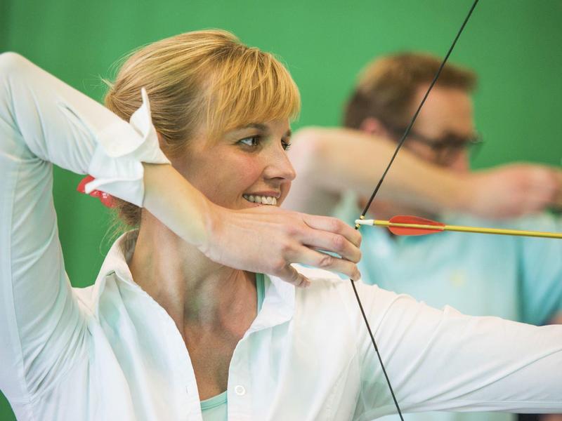 Kobieta trenuje łucznictwo w pomieszczeniu, skupiona z napiętym łukiem.