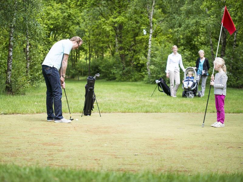 Osoba grająca w golfa na polu z innymi golfistami i wózkami golfowymi w tle.