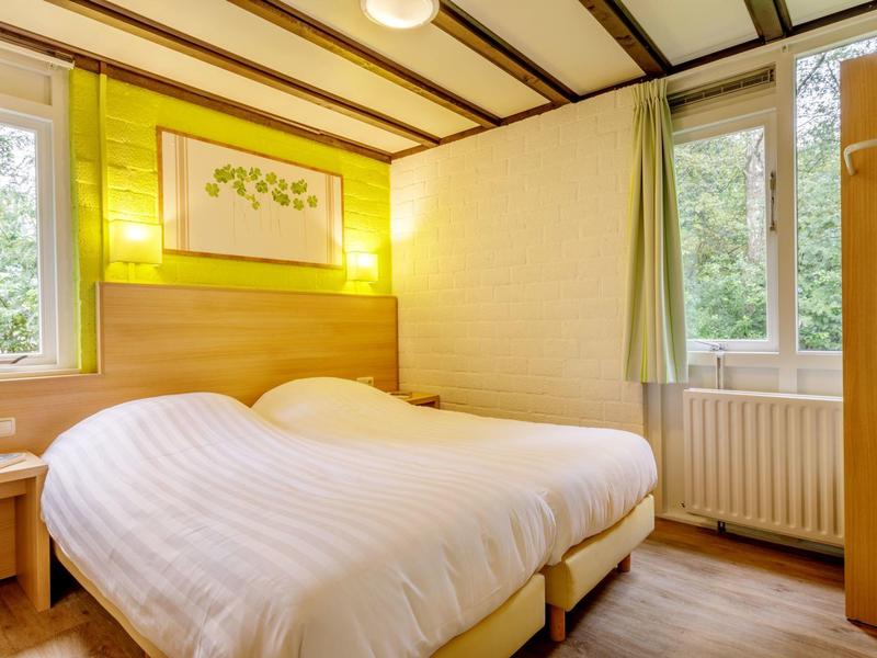 Jasny pokój hotelowy z dwoma pojedynczymi łóżkami, drewnianym sufitem i oknami z widokiem na zieleń.