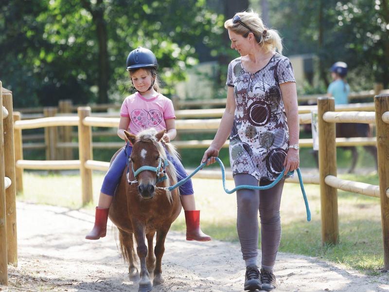 Bambino cavalca un pony su un sentiero all'aperto, guidato da un adulto accanto a una recinzione di legno.