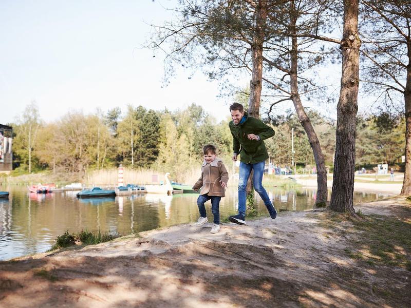 Due persone passeggiano lungo la riva di un lago con barche e alberi sullo sfondo.