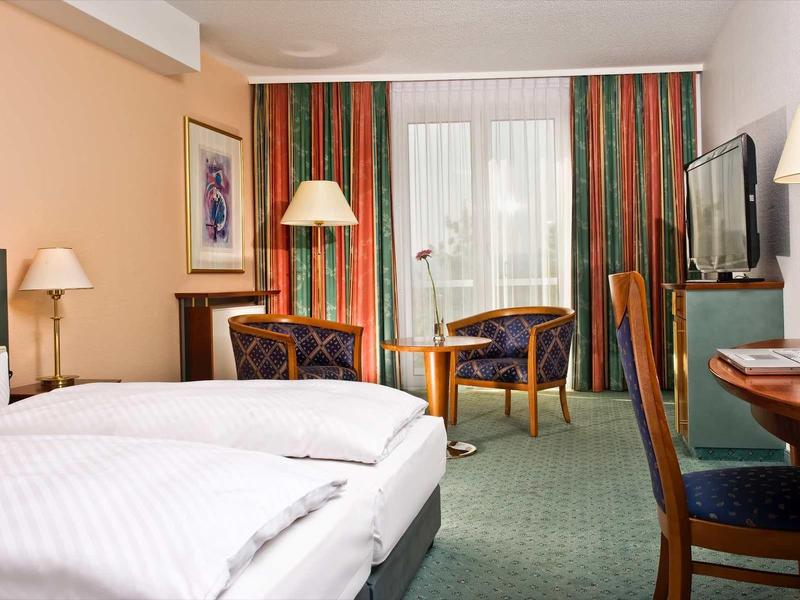 Hell eingerichtetes Hotelzimmer mit Doppelbett, Sesseln, Schreibtisch und großem Fenster mit Gardinen.