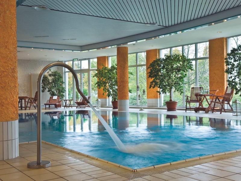 Innenraum mit kleinem Schwimmbecken, Wasserstrahl, Pflanzen und Sitzgelegenheiten am Fenster.