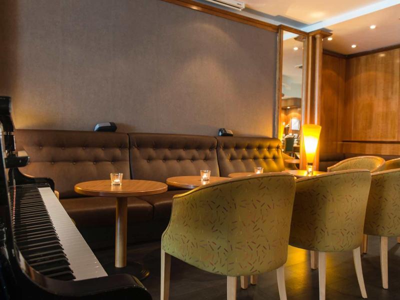 Gemütliche Lounge mit braunem Sofa, gelben Stühlen, Holztischen und Klavier in warmem Licht.