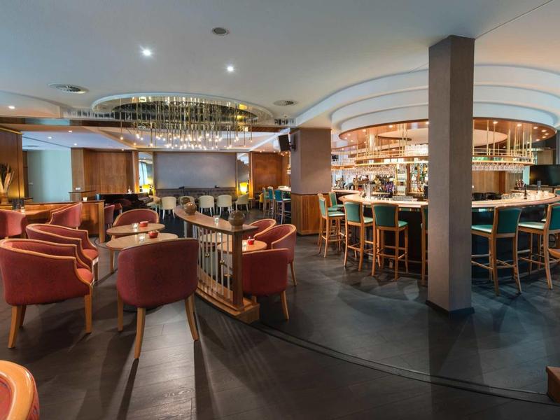 Moderne Bar mit runden Tischen, roten Sesseln, beleuchtetem Tresen und warmem Holzdekor.