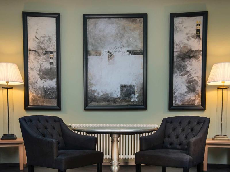 Zwei schwarze Sessel vor Heizkörper mit drei abstrakten Bildern und Stehlampen an grauer Wand.