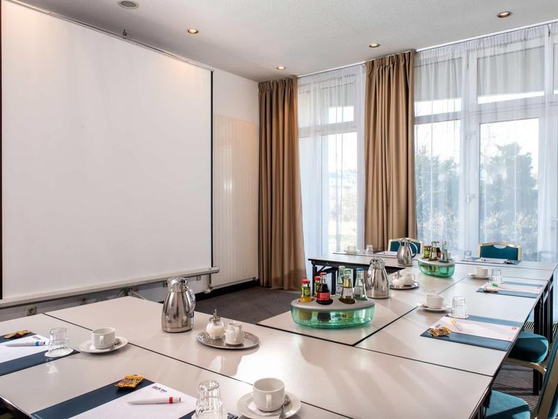 Modernes Konferenzzimmer mit U-förmigem Tisch, Notizblöcken, Tee-Set und großer Leinwand vor Fensterwand.