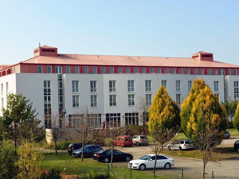 Großes, weißes Gebäude mit rotem Dach, umgeben von Bäumen, Autos und einem Parkplatz.