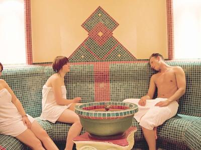 Trzy osoby w białych ręcznikach siedzą w turkusowym spa z mozaiką i fontanną.