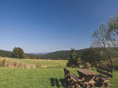 Zielone pole z drewnianym stołem i ławkami pod niebieskim niebem, otoczone wzgórzami.