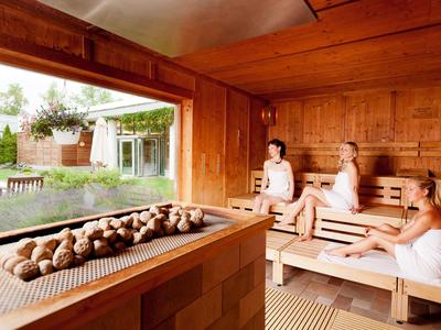 Drei Frauen in weißen Handtüchern sitzen in einer hölzernen Sauna mit integriertem Ofen.