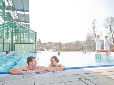 Menschen entspannen im Außenpool eines modernen Wellnessbads bei sonnigem Wetter.