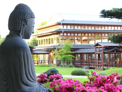 Buddha-Statue im Vordergrund vor einem traditionellen Gebäude mit Blumen im Garten.