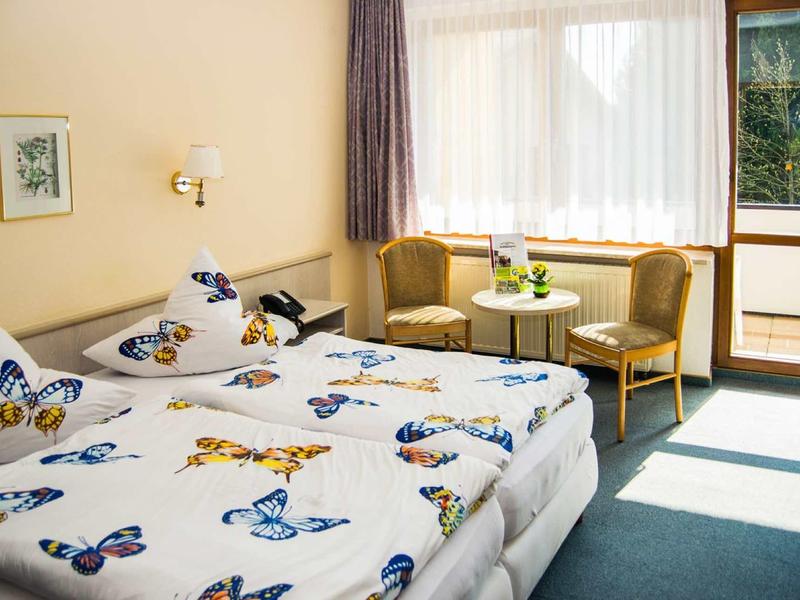 Helles Zimmer mit Doppelbett, Schmetterlingsbettwäsche, zwei Sesseln, kleinem Tisch und großem Fenster.