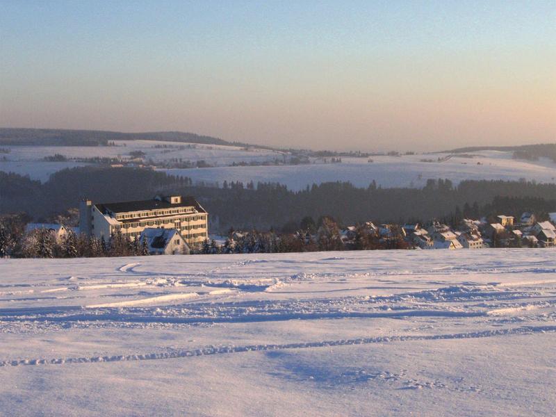 Schneebedeckte Landschaft mit Häusern und einem großen Gebäude im Hintergrund bei Sonnenuntergang.