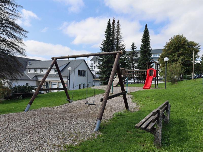 Spielplatz mit Schaukel, roter Rutsche, Holzbänken, Grasfläche und Bäumen im Hintergrund.