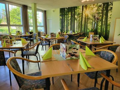 Helles Restaurant mit Holztischen, grünen Servietten und Waldwanddekoration.