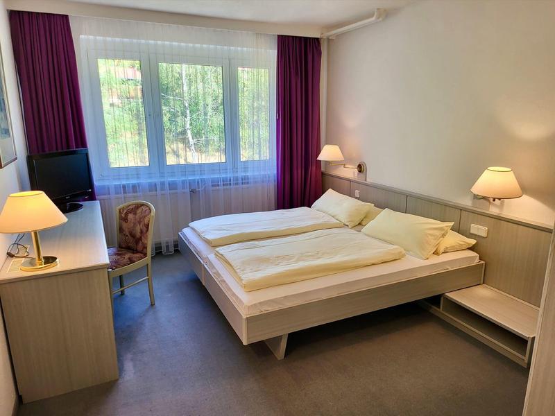 Ein einfach eingerichtetes Schlafzimmer mit Doppelbett, zwei Nachttischlampen und einem Fenster mit lila Vorhängen.