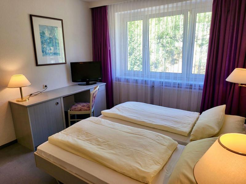 Hell eingerichtetes Schlafzimmer mit zwei Einzelbetten, Schreibtisch, Stuhl und großen Fenstern.