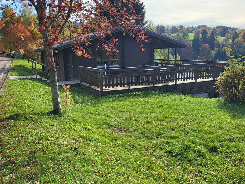 Modernes Holzhaus mit großer Terrasse, umgeben von grünem Rasen und bunten Herbstbäumen.