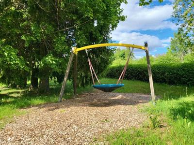 Eine blaue Nestschaukel hängt an einem Holzrahmen in einem grünen Park mit Bäumen und blauem Himmel.