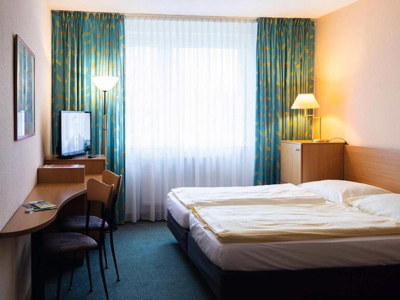 Einfaches Hotelzimmer mit Doppelbett, Schreibtisch, Stühlen, Fernseher und großen grünen Vorhängen.