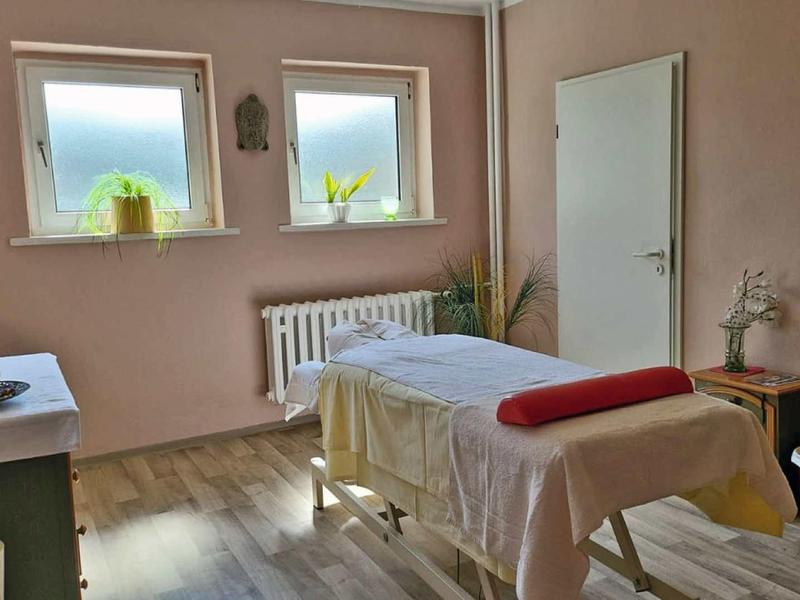Gemütlicher Behandlungsraum mit Massagebank, zwei Fenster, Holzboden und Rosa Wänden.