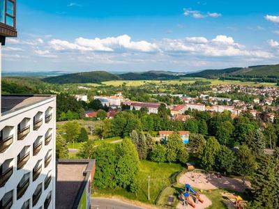 Blick über eine Stadt mit Häusern, viel Grün, Spielplatz und hügeliger Landschaft im Hintergrund.