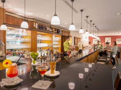 Moderne Hotelbar mit langen Tresen, Stühlen und Getränken, beleuchtet durch hängende Lampen.