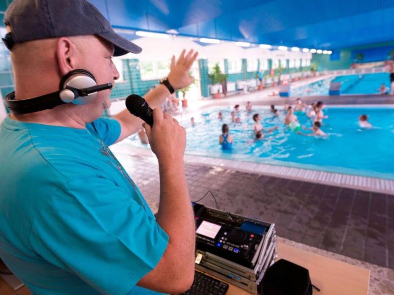 Annonceur avec microphone debout près d'une piscine, des gens dans l'eau.