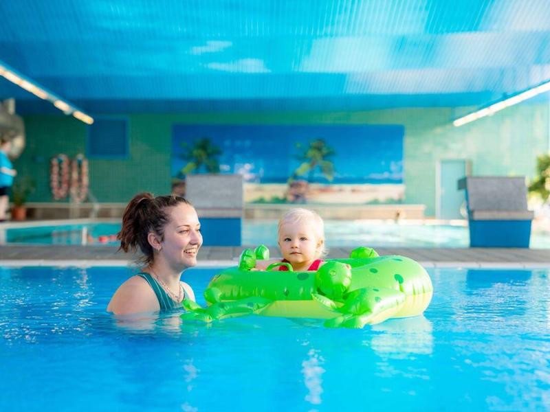 Femme et bébé avec bouée verte dans piscine intérieure, plafond lumineux et fond bleu.