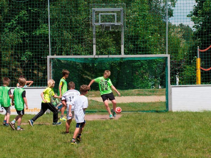 Des enfants jouent au football sur un terrain enherbé devant un but avec filet et un panier de basket en arrière-plan.