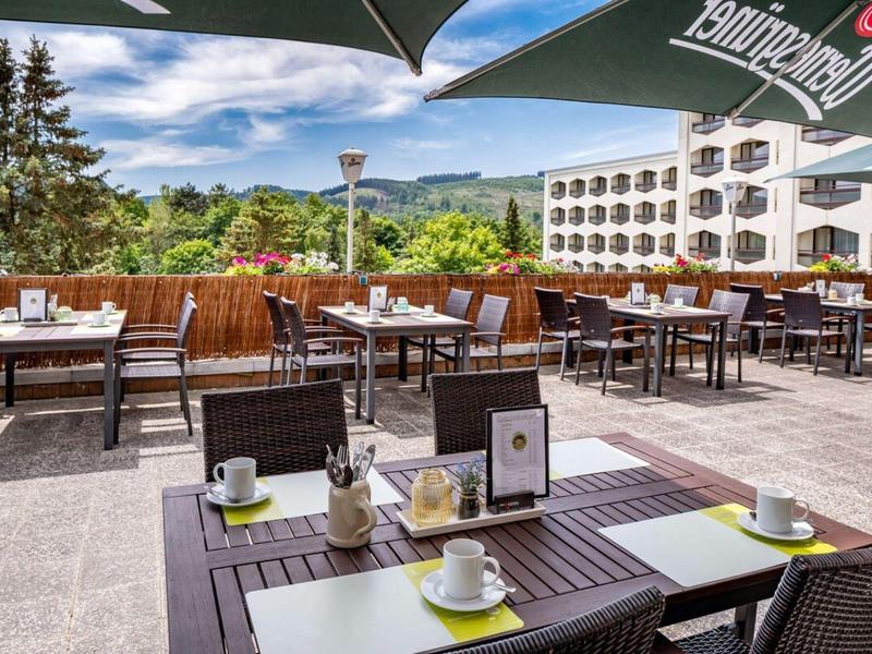 Terrasse d'hôtel avec tables, chaises et vue sur des collines vertes sous un ciel bleu.