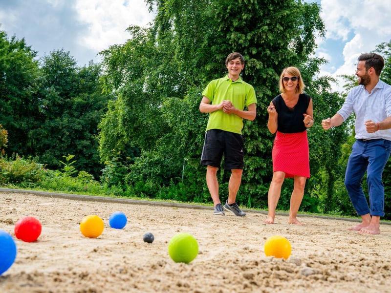 Trois personnes jouent à la pétanque sur un terrain sablonneux en plein air par temps ensoleillé.