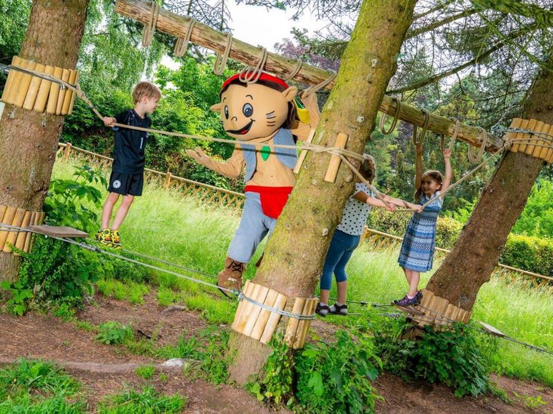 Des enfants grimpent sur un parcours de corde en plein air avec une personne en costume de mascotte coloré.