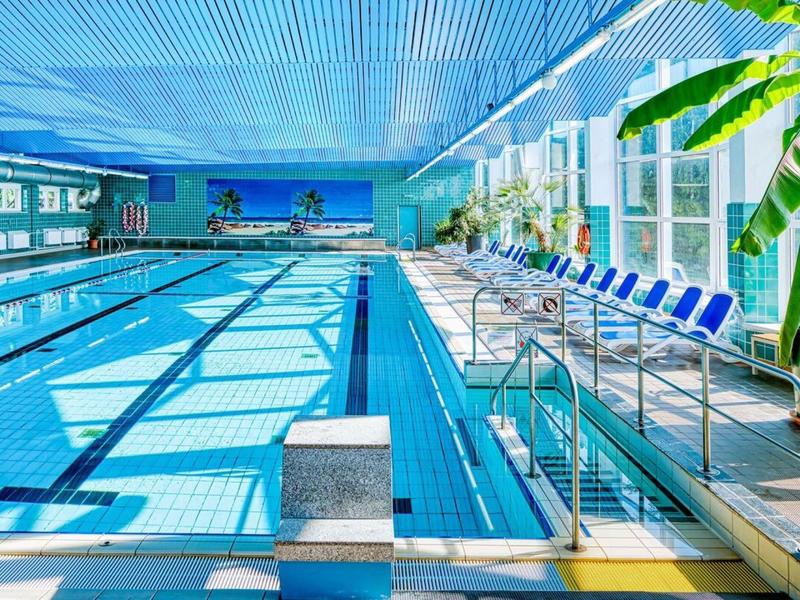 Piscine intérieure avec longues lignes, chaises bleues et grandes plantes à droite.