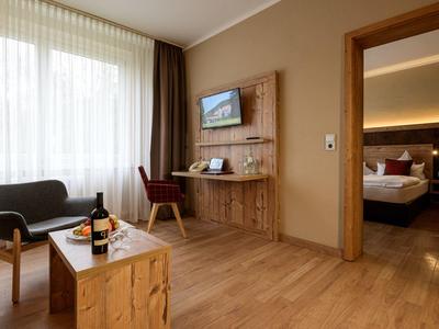 Bright hotel room with living area, wooden floor, seating, workspace, and view into bedroom.
