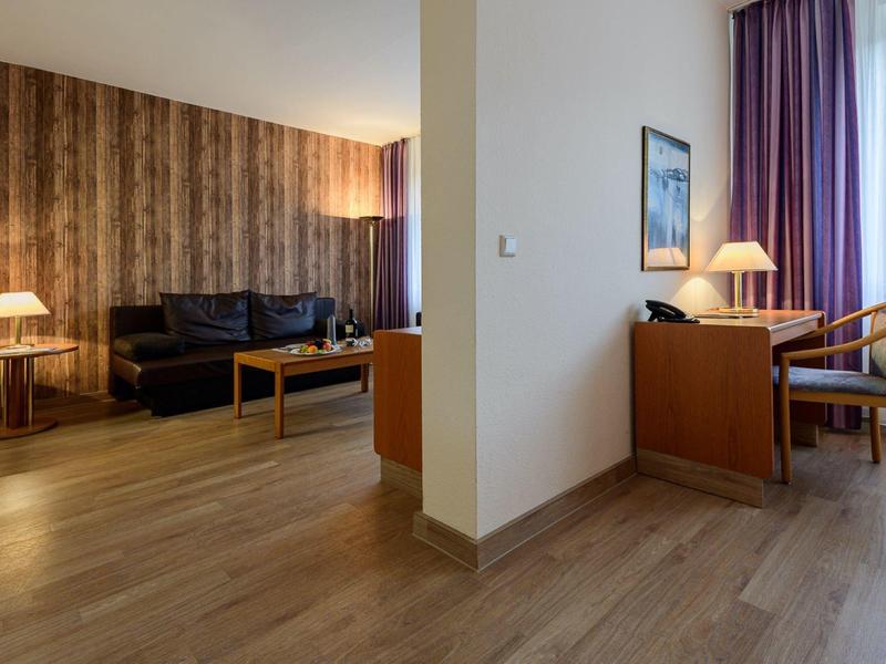 Hotel room with seating area, wooden flooring, floor lamp, and desk with chair beside purple curtains.