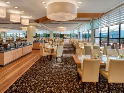 Helles Restaurant mit großen Fenstern, Holzboden und beige Polsterstühlen.