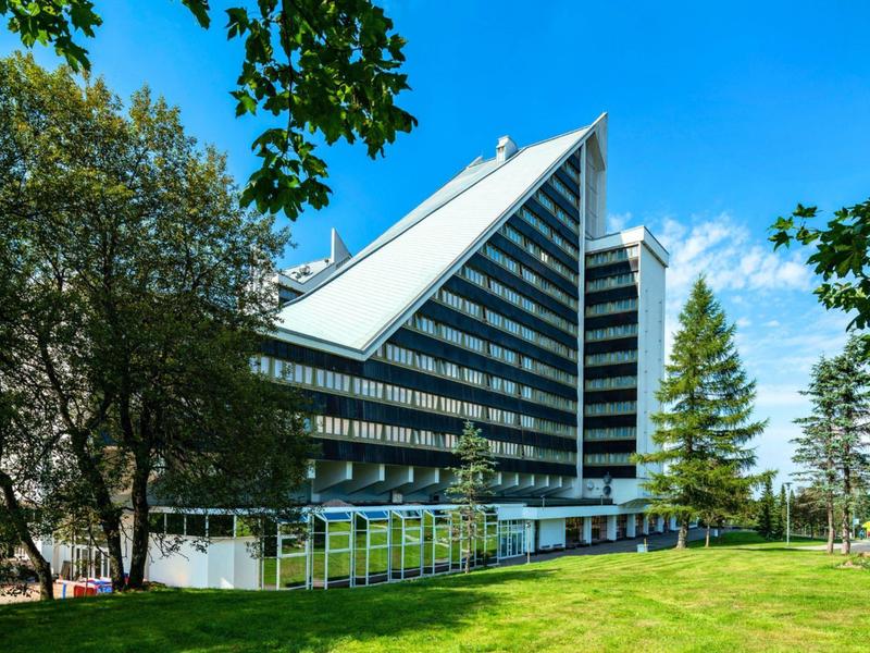 Modernes Hotelgebäude mit schrägem Dach, umgeben von Bäumen und grüner Wiese unter blauem Himmel.