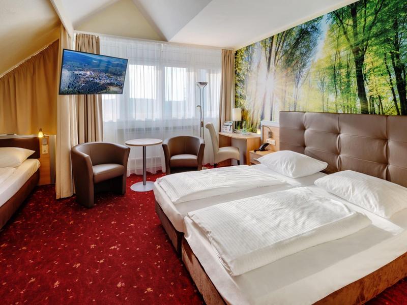Gemütliches Hotelzimmer mit Doppelbett, zwei Sesseln, TV und großer Waldwanddekoration.