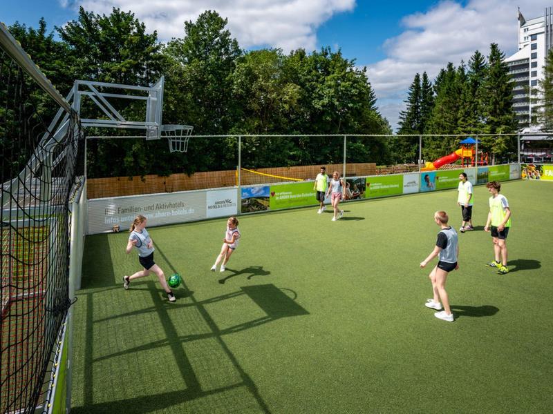 Kinder spielen Fußball auf einem kleinen Rasenspielfeld im Freien bei Tageslicht.